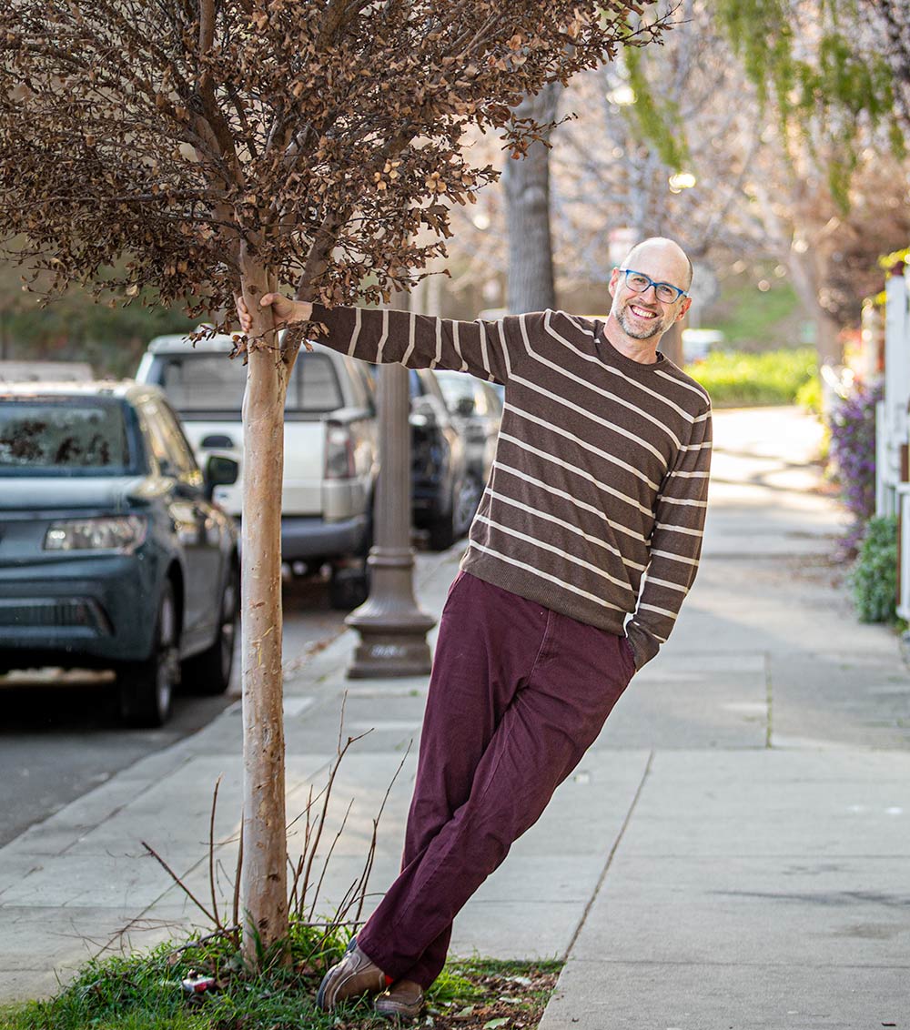 Greg Goodman, founder of Goodman Creatives, leaning against a tree on a neighborhood street